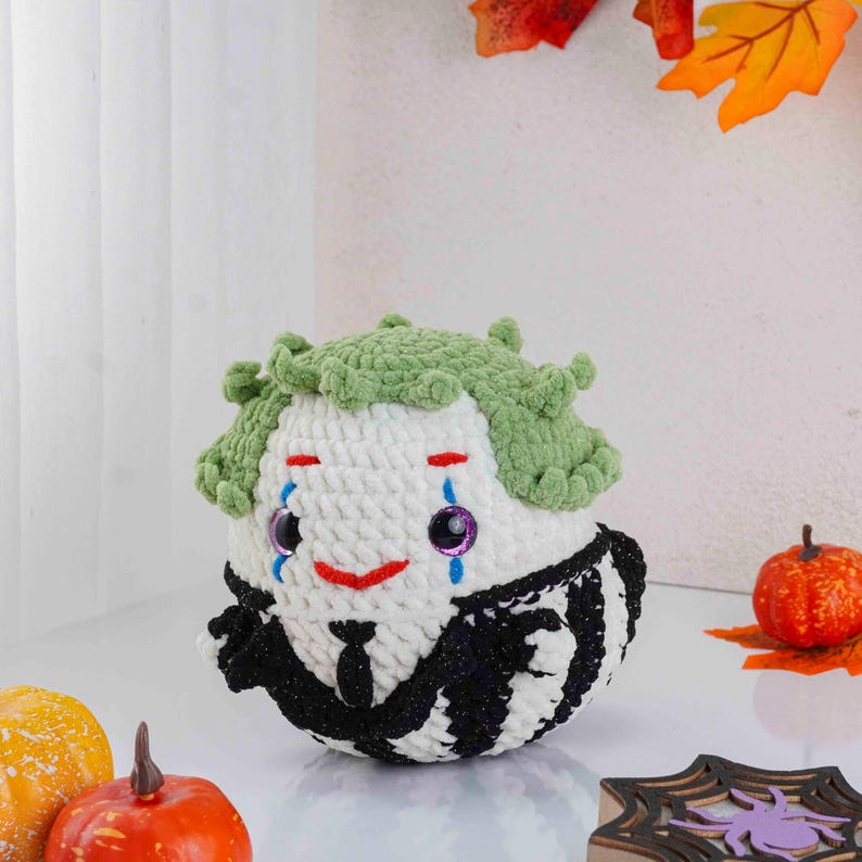 May include: A handmade, round, white and black striped crochet doll with green hair, a red smile, and blue and purple teardrops. The doll has a black suit and tie. The doll is a Halloween or spooky themed item.
