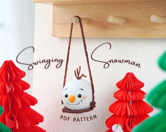 May include: A crocheted snowman with a brown hat and a brown scarf is hanging from a brown rope swing. The snowman is white with black eyes and a carrot nose. The background is a white wall with red and green paper trees. The text "Swinging Snowman PDF Pattern" is written below the snowman.