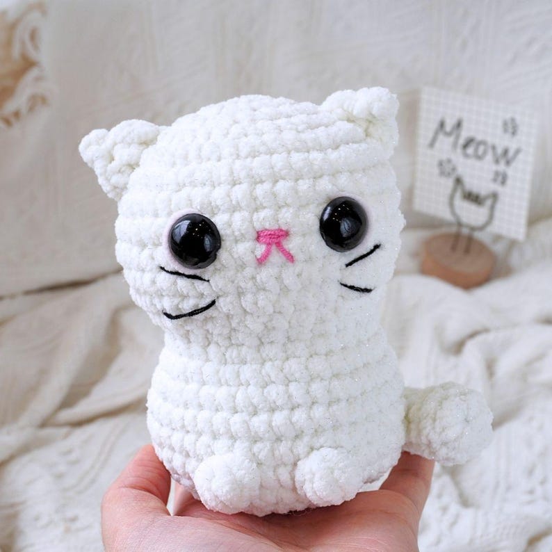 May include: White crocheted cat plush toy with black eyes, a pink nose, and black whiskers. The toy is sitting on a hand.