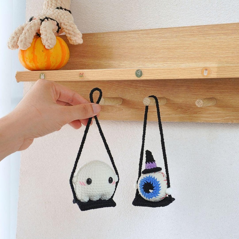 May include: Two crocheted Halloween decorations hanging on a wooden shelf. One is a white ghost with a black swing set and the other is a blue eye with a purple witch hat and a black swing set.
