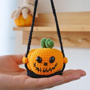 May include: A small crocheted pumpkin with a black strap, black eyes, and a stitched smile. The pumpkin is orange with a green stem.