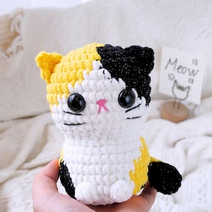 May include: A crocheted stuffed cat toy with black, white, and yellow yarn. The cat has large black eyes and a pink nose.