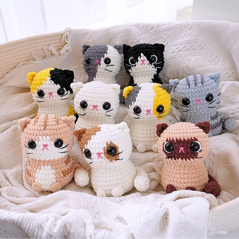 May include: A collection of nine crocheted cat plushies in various colours and patterns, including white, grey, brown, black, and yellow. The plushies have black eyes and pink noses.