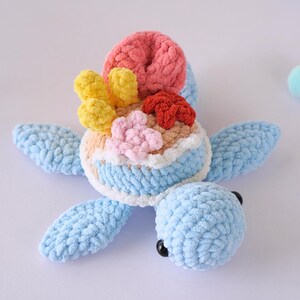 May include: A crocheted blue turtle toy with a shell decorated with colorful coral and flowers. The turtle has black eyes and a light blue body.