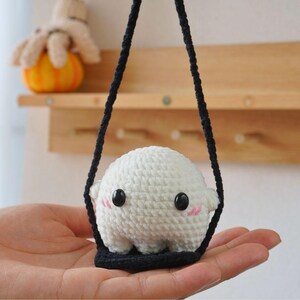 May include: A white crocheted ghost with black eyes and pink cheeks is hanging from a black string. The ghost is sitting on a black crocheted platform.