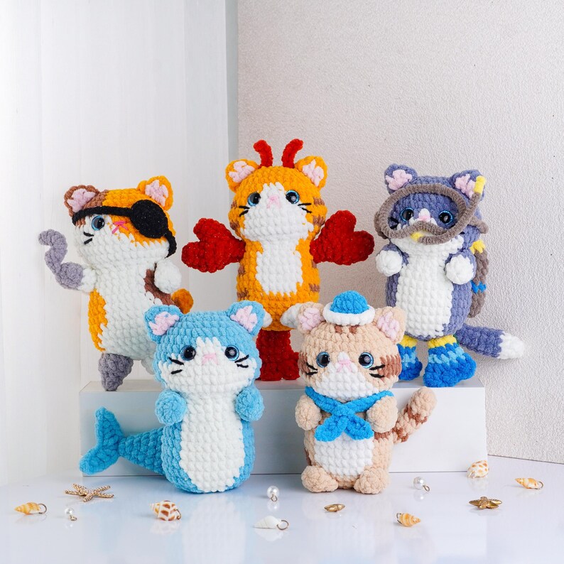 May include: Five handmade crochet cat figurines in various colors and costumes. One cat wears an eye patch, another has wings, and a third has a mermaid tail. The cats are white, orange, blue, and tan.