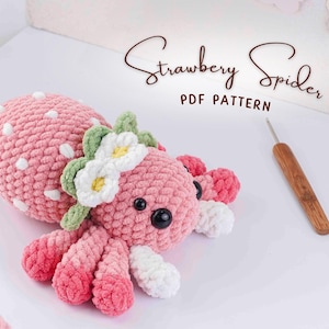 Könnte beinhalten: Handgemachte, rosa gehäkelte Spinne mit weißen Tupfen, einem grünen und weißen Blumenkranz und schwarzen Augen. Der Text "Strawberry Spider PDF Pattern" ist in einer Schreibschrift geschrieben. Ein Häkelhaken und eine Schere sind ebenfalls sichtbar.