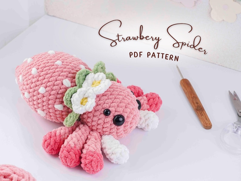 May include: A handmade pink crocheted spider with white polka dots, a green and white flower crown, and black eyes. The text "Strawberry Spider PDF Pattern" is written in a script font. A crochet hook and scissors are also visible.