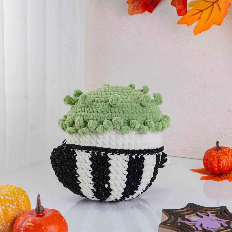 May include: A handmade crochet pumpkin-shaped container. The container has a black and white striped base, a white midsection, and a green top with textured details. The container is decorated with small orange pumpkins and fall leaves.