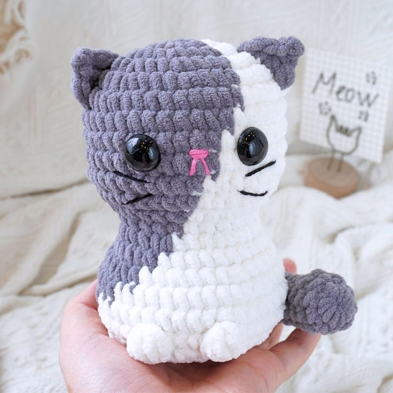 May include: A grey and white crocheted cat plush toy with black eyes and a pink nose. The toy is sitting in a person's hand. The background is a white blanket with the word "Meow" written on a sign.
