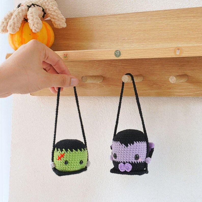 May include: Two crocheted Halloween characters hanging from hooks on a wooden shelf. The character on the left is green and has a stitched-on scar. The character on the right is purple and has a black bow tie.