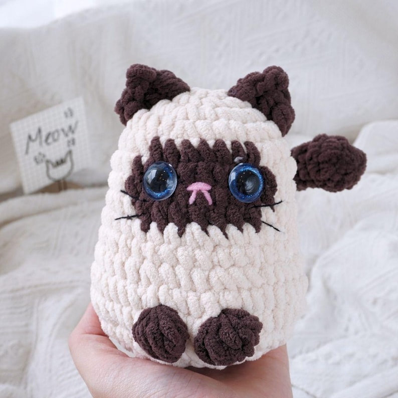 May include: A crocheted stuffed cat toy with brown ears, paws, and a face. The cat has blue eyes and a pink nose. The cat is sitting on a hand.