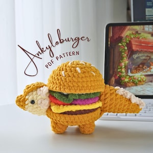 Op de afbeelding: Een handgemaakt gehaakt pluche speelgoed in de vorm van een hamburger met dinosauruskenmerken. Het speelgoed is voornamelijk geel, met groene, roze en bruine lagen. De tekst "Ankyloburger PDF PATTERN" is cursief geschreven.