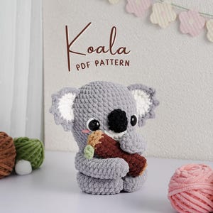 May include: A crocheted gray koala bear holding a brown log. The koala has a black nose, large eyes, and white-lined ears. The text "Koala PDF PATTERN" is above the koala. Yarn balls in brown, green, pink, and white are in the background.
