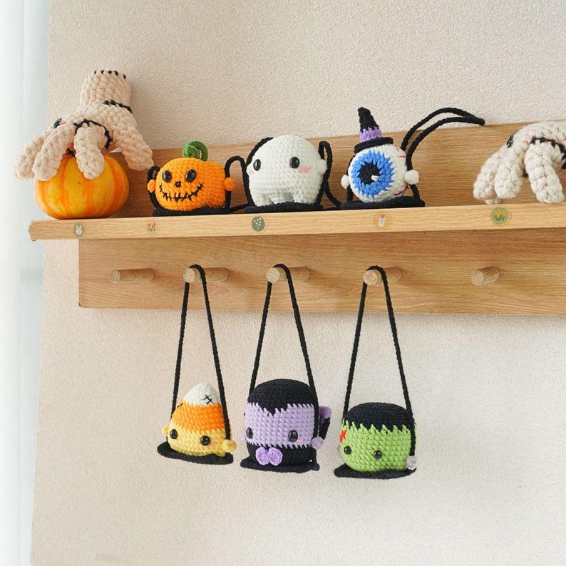 May include: A wooden shelf with hooks holds a variety of Halloween-themed crocheted toys. The toys include a pumpkin, a ghost, an eye, a candy corn, a vampire, and a Frankenstein.