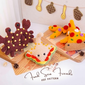 May include: Three crocheted plush toys: a dark brown crab with yellow spots, a beige sandwich with red and green layers, and a yellow pizza slice with red pepperoni and purple toppings. The text "Food Sea Friends PDF PATTERN" is visible below.
