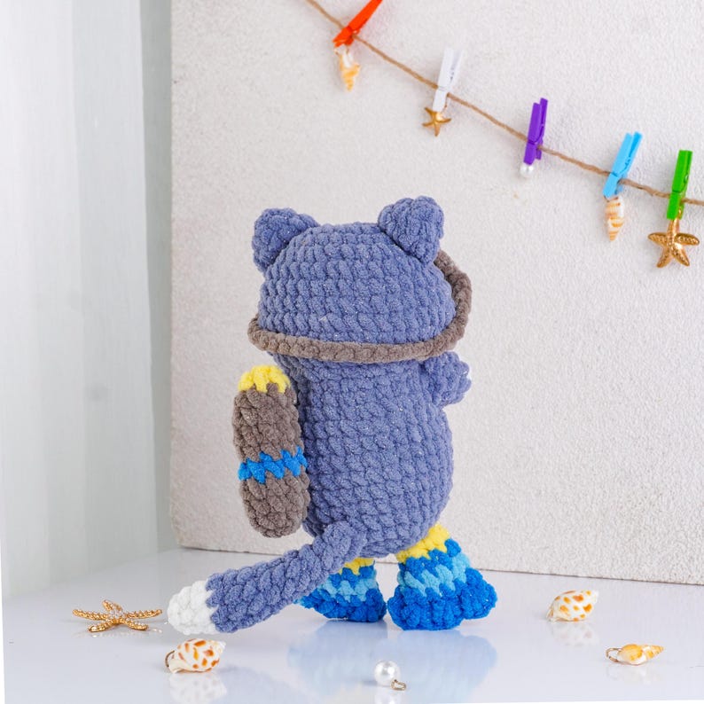 May include: A handmade blue crochet cat doll with a brown and yellow accessory. The cat has a white tail tip and blue and yellow accents on its feet. The doll is standing on a white surface with decorative seashells and starfish.
