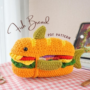 May include: A crocheted fish bread amigurumi toy. The toy is shaped like a fish and has a bread-like filling. The toy is made with a variety of colors, including yellow, orange, green, and red. The toy is sitting on a pink and white checkered surface. The text "Fish Bread PDF Pattern" is visible in the image.