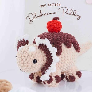 May include: A crocheted dinosaur toy in beige, brown, and white, topped with a red cherry. The dinosaur has a dessert-like appearance, with a brown, textured topping. The text "PDF PATTERN Dilophosaurus Pudding" is visible.