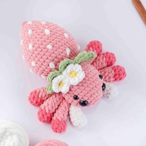 May include: A handmade pink crochet strawberry octopus with white polka dots, green leaves, and white flowers with yellow centers. The octopus has eight pink and white tentacles and black eyes.