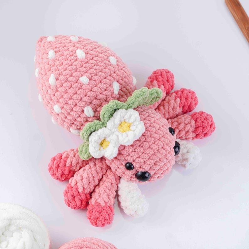 May include: A handmade pink crochet strawberry octopus with white polka dots, green leaves, and white flowers with yellow centers. The octopus has eight pink and white tentacles and black eyes.