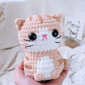 May include: A crocheted plush toy cat with orange and white fur. The cat has large black eyes, a pink nose, and a white face. The cat is sitting on a hand.