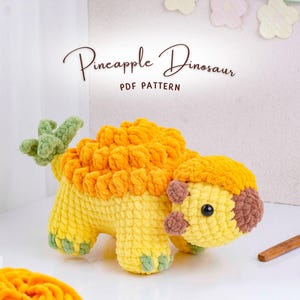 May include: A crocheted Pineapple Dinosaur in yellow, orange, and green. The plush toy has a textured body, a spiky back, and a small head with brown accents. The text "Pineapple Dinosaur PDF Pattern" is above the toy.