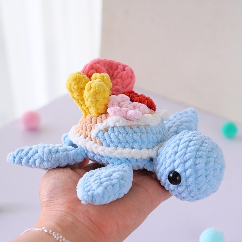 May include: A crocheted blue sea turtle with a colorful coral reef on its back. The turtle has black eyes and a white shell with a light blue trim.