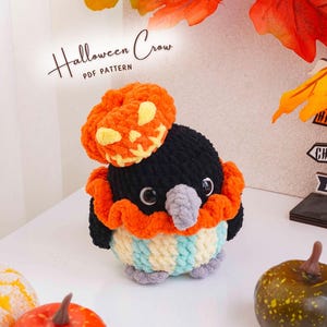 Può includere: Un peluche a forma di corvo realizzato all'uncinetto con testa nera, becco grigio e corpo a righe azzurre e bianche. Il corvo indossa un colletto arancione a balze e un piccolo cappello a forma di zucca. Testo: Halloween Crow PDF PATTERN.