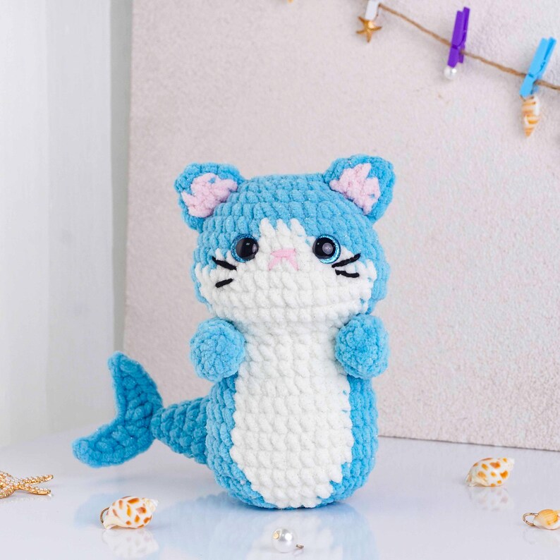 May include: A handmade blue and white crochet cat-mermaid plush toy. The plush has a blue head with pink inner ears, black whiskers, and large black eyes. The body is white with blue arms and a blue tail. Seashells are scattered around the toy.