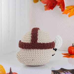 May include: A handmade, beige crocheted acorn with a brown band and cross-shaped detail on top. The acorn has a white, leaf-shaped detail and a small, black face. The background includes fall foliage and a small pumpkin, suggesting a seasonal theme.