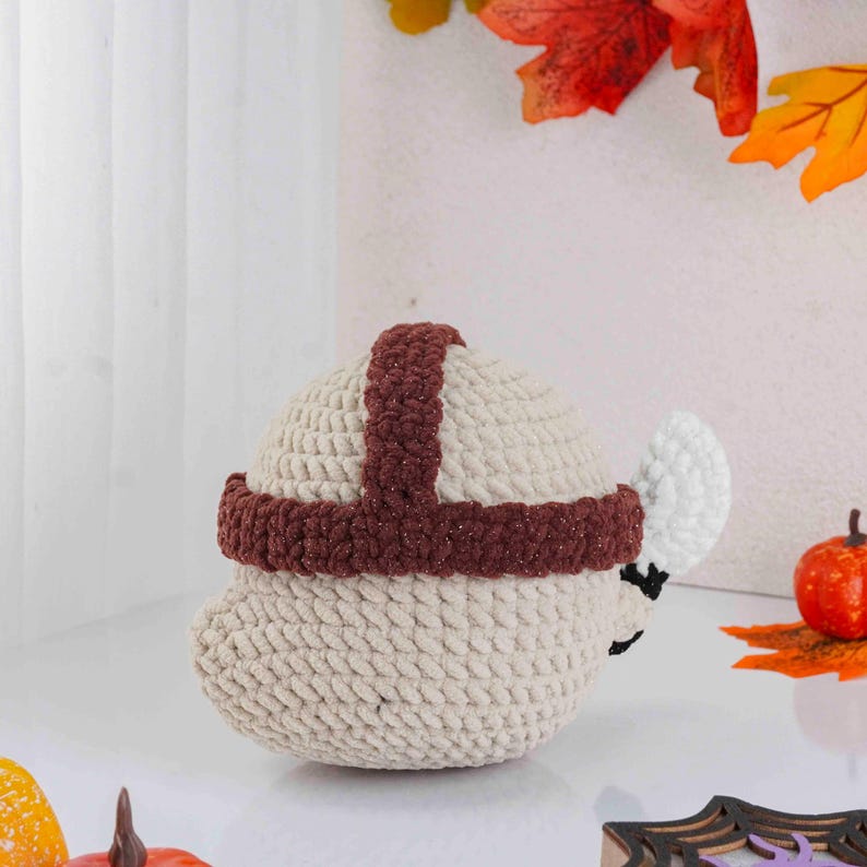 May include: A handmade, beige crocheted acorn with a brown band and cross-shaped detail on top. The acorn has a white, leaf-shaped detail and a small, black face. The background includes fall foliage and a small pumpkin, suggesting a seasonal theme.