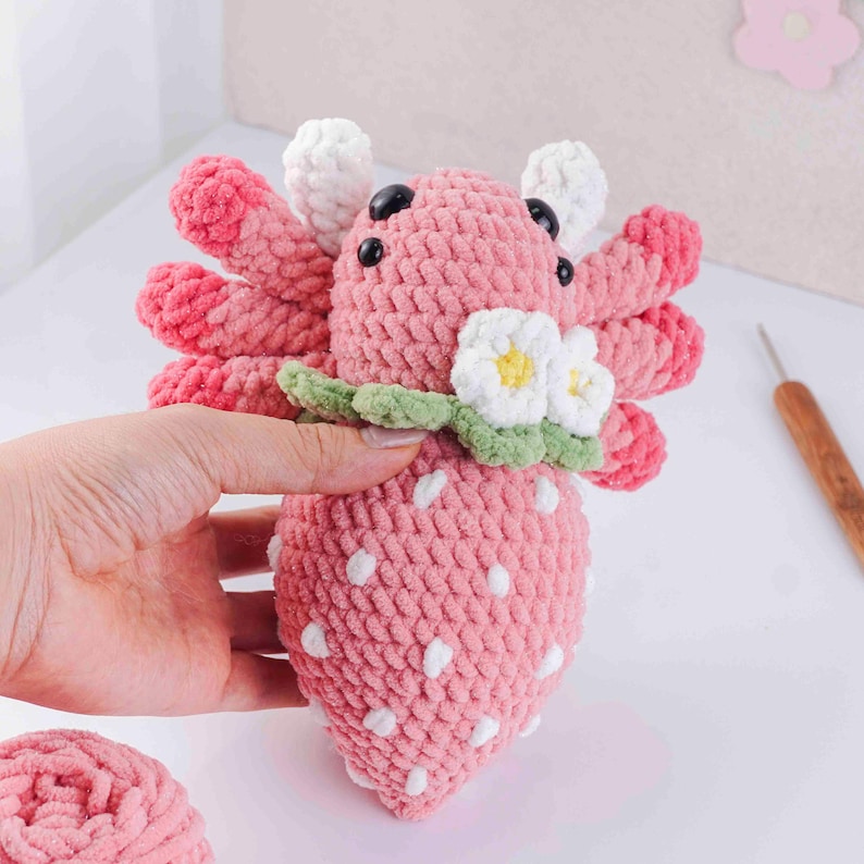 May include: A handmade crochet axolotl plushie in pink, resembling a strawberry. The plushie features white spots, black eyes, and white and pink gills. It has a green leaf collar with white and yellow flower accents. A ball of pink yarn is in the foreground.