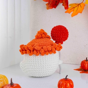 May include: A handmade, white and orange crochet pumpkin decoration. The pumpkin has a white base, an orange top, and a red lollipop-like accent. The decoration is likely for the fall season or Halloween.