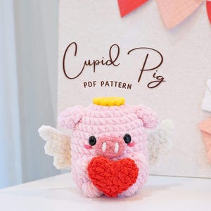 Puede incluir: Un cerdo de crochet rosa con un halo amarillo y alas de ángel blancas, sosteniendo un corazón rojo. El cerdo es un patrón de crochet llamado "Cupid Pig" y está disponible para descargar en PDF.