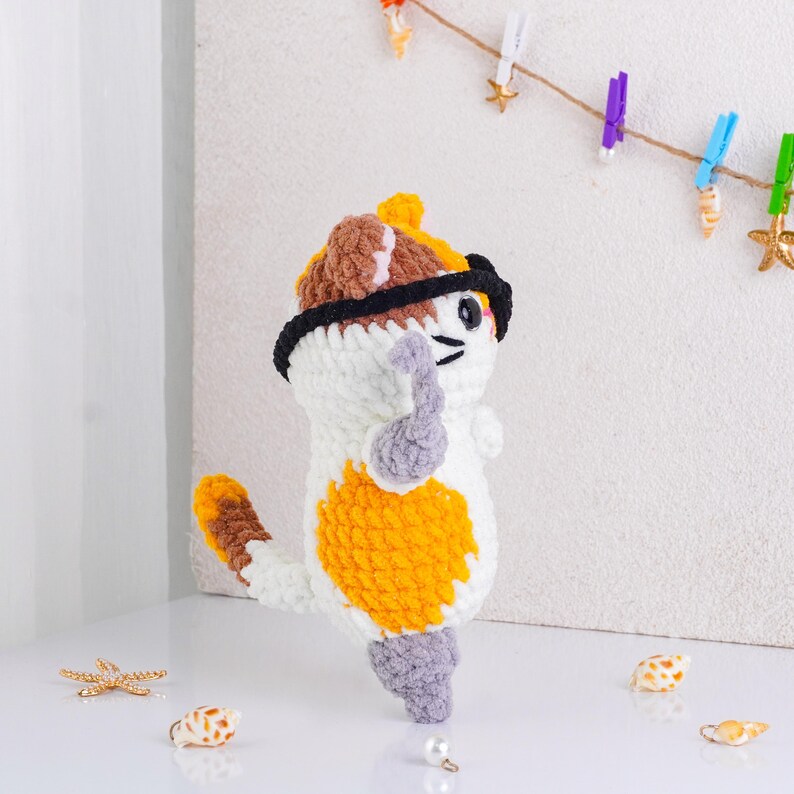 May include: A handmade crochet cat doll with a white, orange, brown, and gray color scheme. The cat wears a black hat and glasses. The cat is standing upright with its arm raised. Seashells and a pearl are in the foreground.