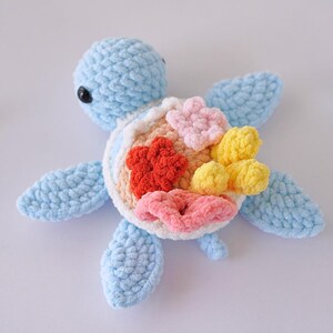 May include: A crocheted blue sea turtle with a colorful coral reef design on its shell. The coral is made of pink, yellow, and orange yarn.