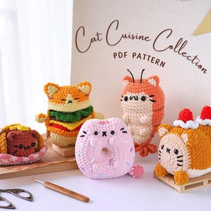 Op de afbeelding: Een verzameling gehaakte katten-thema voedsel speelgoed. De speelgoed zijn gevormd als een hamburger, een donut, een garnaal en een taart. De speelgoed zijn allemaal gemaakt met verschillende kleuren garen en hebben schattige kattengezichten.