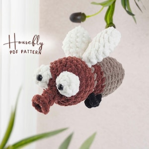 May include: A crocheted housefly toy with a brown body, white wings, and large white eyes. The fly has a long, textured proboscis and small black feet. The text "Housefly PDF Pattern" is in the upper left corner.