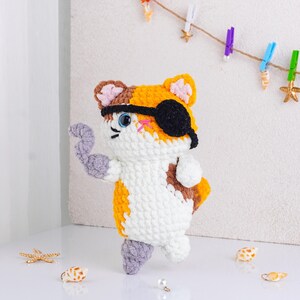 May include: A handmade crochet cat toy in white, orange, brown, and gray. The cat has a black eye patch and is posed with one arm raised. The toy is decorated with blue eyes and pink cheeks. The cat is standing on a white surface.