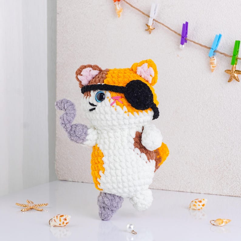 May include: A handmade crochet cat toy in white, orange, brown, and gray. The cat has a black eye patch and is posed with one arm raised. The toy is decorated with blue eyes and pink cheeks. The cat is standing on a white surface.