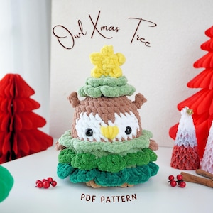May include: A crocheted owl Christmas tree ornament with a brown owl face and a green and brown tree body. The owl has a yellow star on top of its head. The ornament is surrounded by red and white paper Christmas trees.