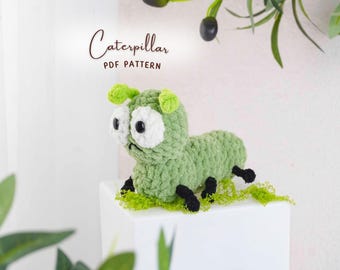 May include: A handmade green crochet caterpillar with white eyes and black pupils, black feet, and green antennae. The caterpillar is sitting on a white surface with green moss. The text "Caterpillar PDF Pattern" is above the caterpillar.