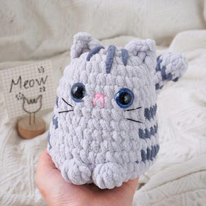 May include: A gray crocheted cat plush toy with blue eyes and a pink nose. The cat is sitting with its paws tucked in front of it. The word "Meow" is written on a small sign behind the cat.