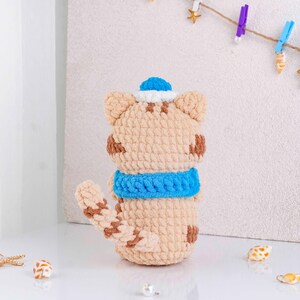 May include: A handmade crochet cat doll in tan with brown spots, a blue scarf, and a blue and white sailor hat. The cat has a long tail and is sitting upright. The doll is made of soft yarn.