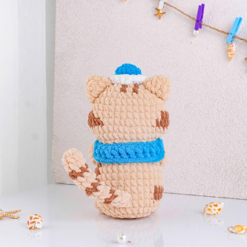 May include: A handmade crochet cat doll in tan with brown spots, a blue scarf, and a blue and white sailor hat. The cat has a long tail and is sitting upright. The doll is made of soft yarn.