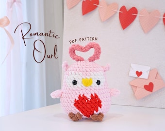 May include: A pink crocheted owl with a red heart on its chest and a pink heart-shaped halo. The owl is sitting on a white surface with a garland of pink and red hearts in the background. The text "Romantic Owl" and "PDF Pattern" is visible in the image.