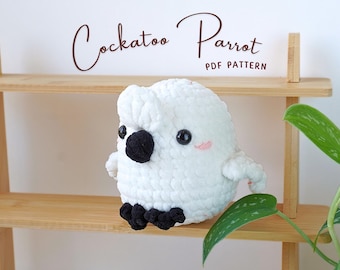 Op de afbeelding: Witte gehaakte kaketoe papegaai pluche speelgoed met zwarte ogen en snavel. Het speelgoed zit op een houten plank met een groene plant op de achtergrond. De tekst "Cockatoo Parrot PDF Pattern" is zichtbaar boven het speelgoed.