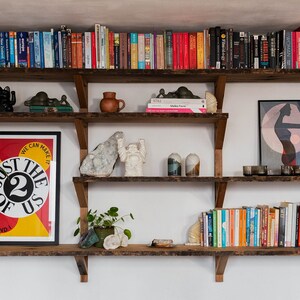 May include: A wooden shelf unit with five shelves, filled with books, plants, and decorative objects. The top shelf has a row of books, the second shelf has a bowl, two black cat figurines, and a green ceramic figurine. The third shelf has a framed poster with the text "Just the 2 of us, we can make it", the fourth shelf has a white ceramic figurine, a grey rock, and two white ceramic containers, and the bottom shelf has a row of books.