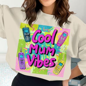 May include: Cream-colored sweatshirt featuring a colorful graphic with the text "Cool Mum Vibes" in pink and purple. The design includes illustrations of retro cell phones in blue, pink, purple, and yellow, set against a vibrant background.